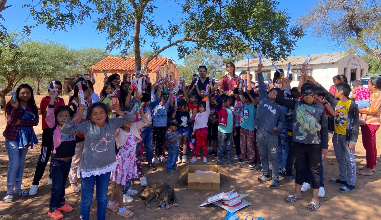 Some forty children stand under trees on bare ground holding toothpaste and toothbrushes in the air, an open box at their feet.